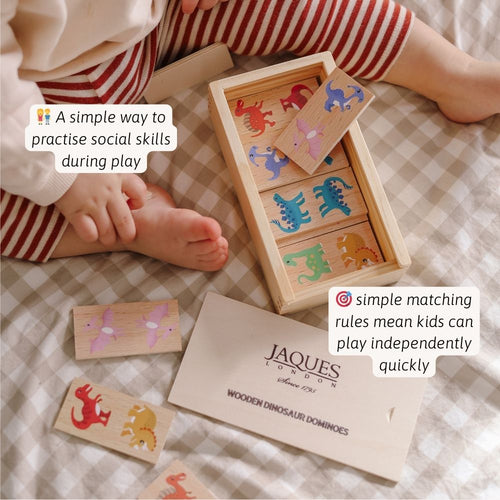 Children playing with wooden dinosaur dominoes set on a checked blanket, with Jaques of London branding. 