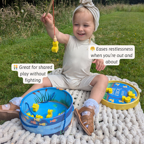 Smiling toddler sitting on a rug playing with the blue duck fishing pond game, holding a magnetic fishing rod with ducks floating inside 
