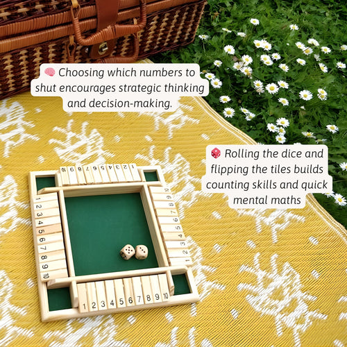 Wooden shut-the-box dice game set on a yellow picnic blanket outdoors, with white flowers in the background and text highlighting strategic thinking, decision-making, counting skills, and quick mental maths. 