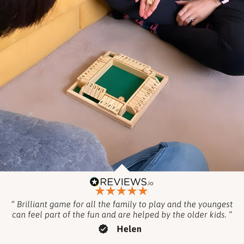 Wooden shut-the-box game placed on the floor between seated players, shown during gameplay with a customer review overlay praising it as a brilliant family game. 