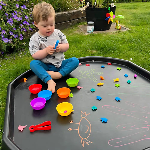 Child playing with colourful toys on a black tray outdoors 