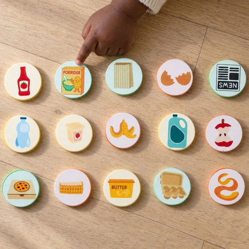 A child's hand points to one of 15 round wooden toy food icons arranged in three rows on a light wood floor. The colorful wooden tokens include illustrations of a red bottle, porridge, pasta, eggshells, a newspaper, a water bottle, a cup, bananas, a milk jug, an apple, a pizza, a basket, butter, eggs.