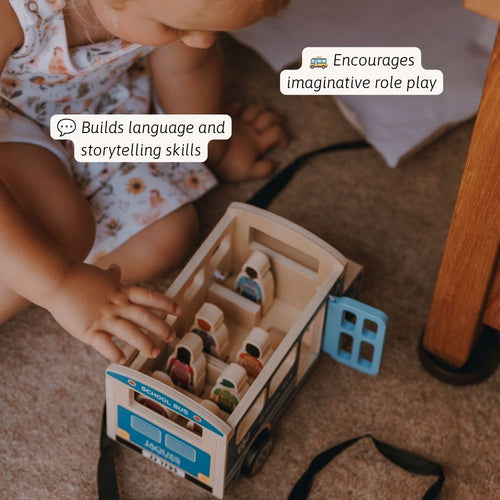 A baby sitting on a carpeted floor playing with a wooden school bus toy. The toy bus has colorful painted windows and small character figures inside. 