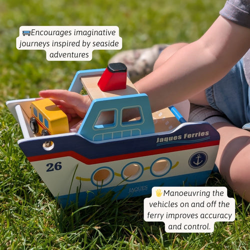 Child’s hand pushing a colorful wooden toy boat across grass outdoors, showing the red roof, blue cabin, yellow smokestack, and painted lifebuoy details on the side. 