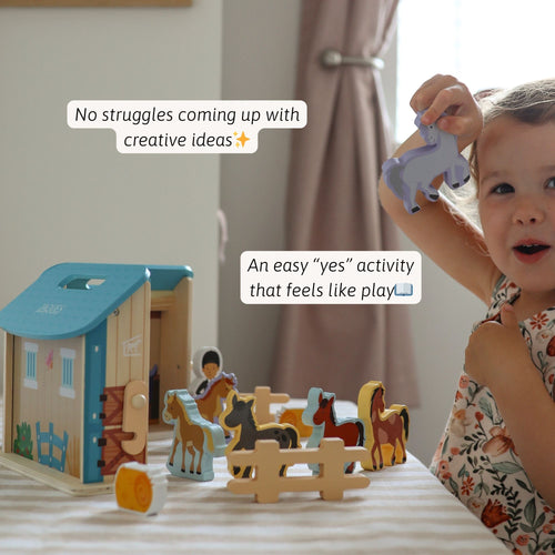Young child smiling while playing with the wooden horse stable and horse figures, suggesting creative and imaginative play. 