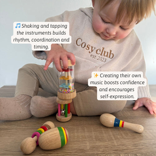 Toddler sitting on the floor playing with a wooden rainmaker and maraca-style rattles, with text about building rhythm, coordination, timing, and confidence through music play. 