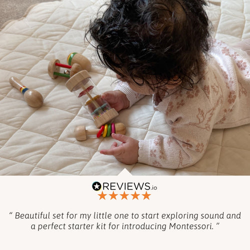 Young child on a quilted mat exploring wooden rattles and bead shakers spread around, with a five-star customer review about introducing sound and Montessori learning. 