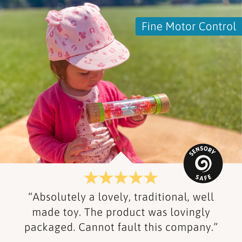 Young child sitting on a picnic blanket examining the rain sound maker, with a fine motor control label and a customer review underneath 