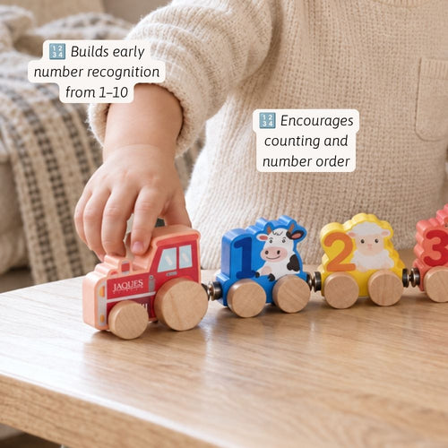 A child's hand plays with a wooden toy train on a light wood table. The train has a pink cab and colorful animal and number carriages, including a blue cow carriage marked '1', a yellow sheep carriage marked '2', and a red bear carriage marked '3'.