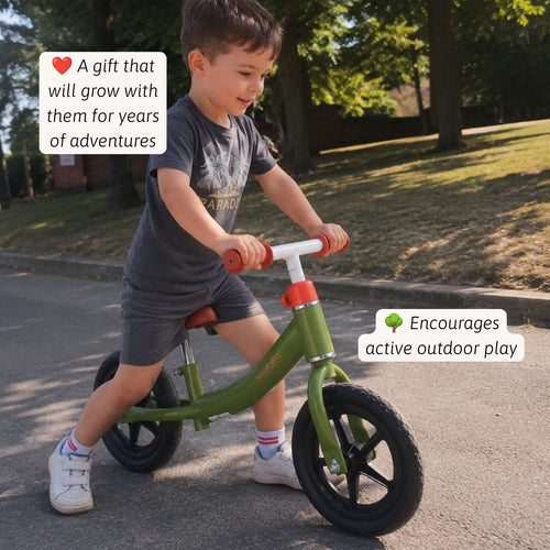 A young boy riding an olive green balance bike outdoors on a paved path. The bike has a white handlebar and red accents. 