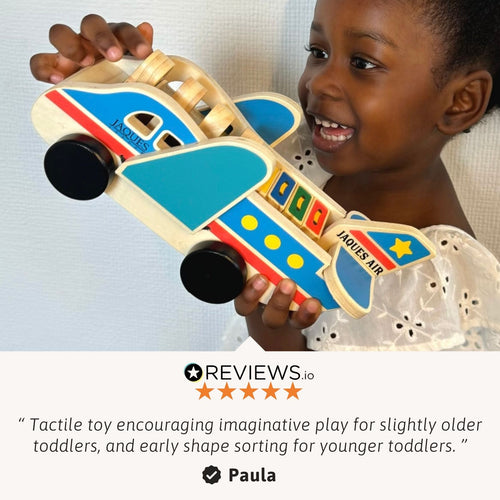 Smiling young child holding a colorful wooden toy airplane with blue wings and orange accents, shown against a light background with small promotional text on the left side. 