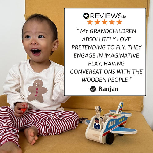 Toddler sitting on the floor holding the wooden airplane next to a product information sign, looking surprised with mouth open in excitement. 