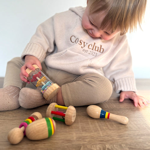 Child playing with wooden toys on a wooden floor, wearing a 'Cosy Club' sweatshirt. 