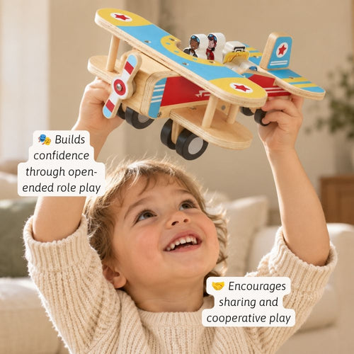A smiling young child in a cream knit sweater holds a colourful wooden bi-plane toy above their head in a bright indoor room. 